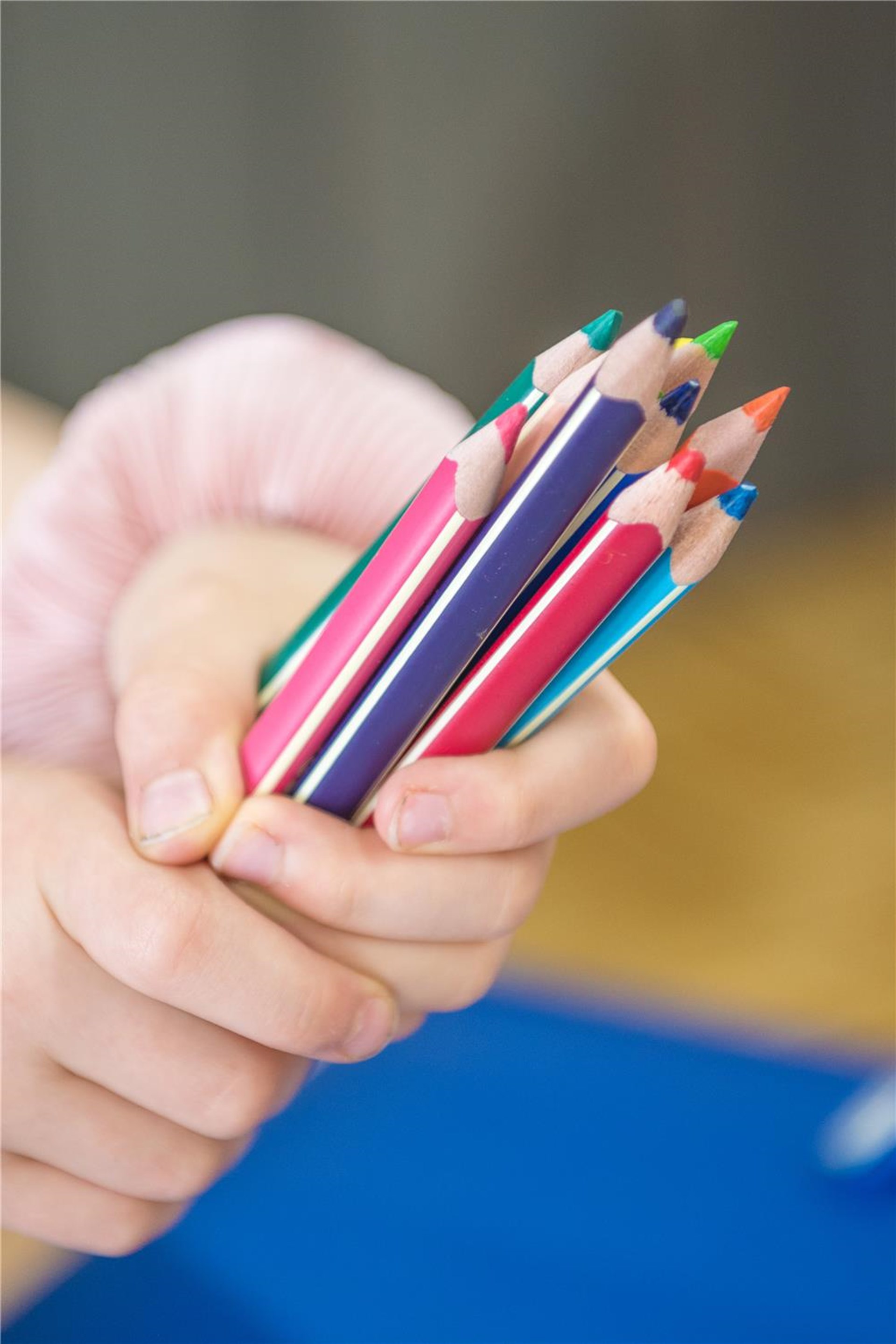 Eine Hand hält mehrere bunte Buntstifte mit gespitzten Spitzen fest zusammen. Im Hintergrund sind ein blaues Blatt Papier und eine verschwommene Umgebung zu sehen.