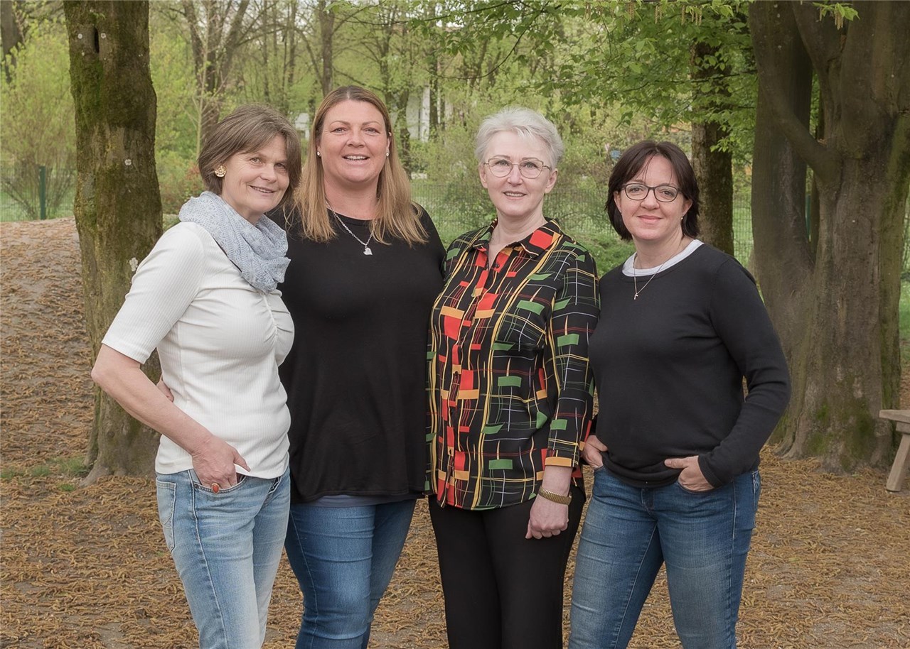 Vier Frauen stehen vor Bäumen und lächeln, umgeben von einer natürlichen Umgebung.