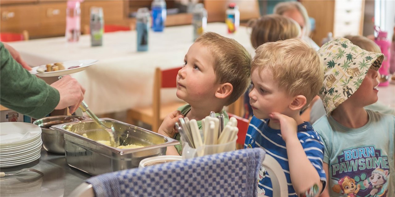 Mehrere Kinder stehen in einer Schlange und warten gespannt auf ihr Mittagessen, das von einem Erwachsenen serviert wird. Im Hintergrund sind Tische mit Wasserflaschen zu sehen
