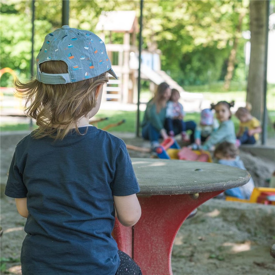 Ein Kind sitzt auf einem Tisch auf einem Spielplatz und schaut den anderen Kindern beim Spielen zu.