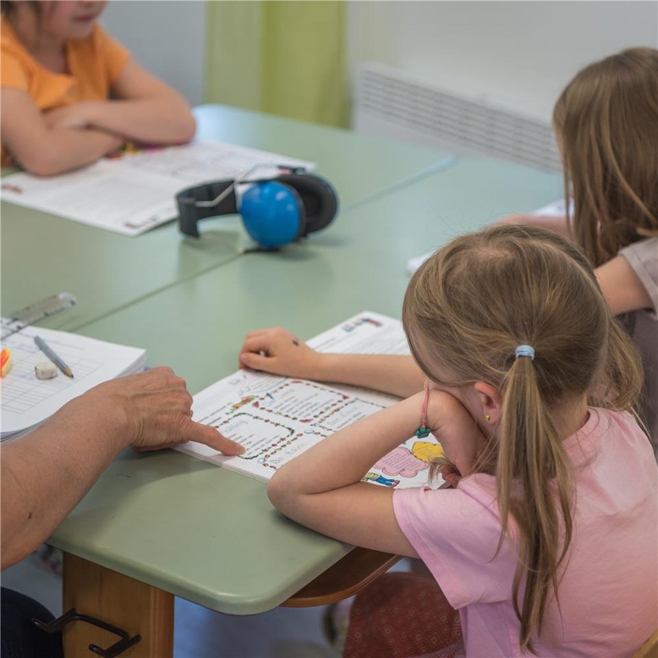 Mehrere Kinder sitzen an einem Tisch und arbeiten an Arbeitsblättern, während eine erwachsene Hand auf ein Blatt zeigt. Auf dem Tisch liegen Schreibutensilien, ein Kopfhörer und weitere Unterlagen.